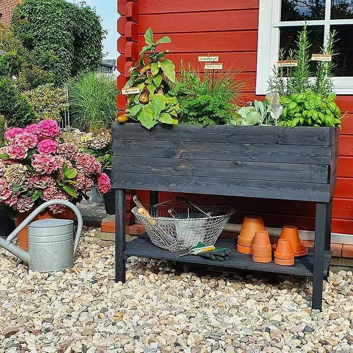 Promadino Hochbeet - 100 x 50 x 80 cm, Holz, Anthrazit - Garten-Hochbeet aus dunklem Holz mit Kräutern, umgeben von Blumen und Gartengeräten, ideal für den Anbau auf kleinem Raum.