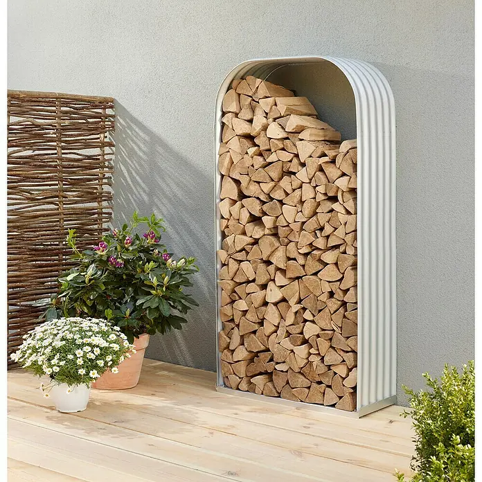 Vitavia Kaminholzregal Lignum 401 - 40 x 99 x 200 cm, Stahl, Blank - Ein moderner, silberfarbener Kaminholzständer, gefüllt mit Holzscheiten, steht auf einer Holzterrasse neben Pflanzen.