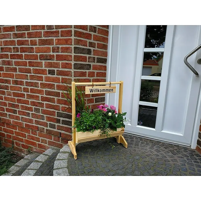 Promadino Pflanzkasten Willkommen - Außenmaß (L x B x H): 59 x 30 x 73 cm, Holz, Braun - Ein hölzerner Blumenkasten mit einem "Willkommen"-Schild, der mit bunten Blumen bepflanzt ist, steht vor einer Backsteinmauer und einer Tür.