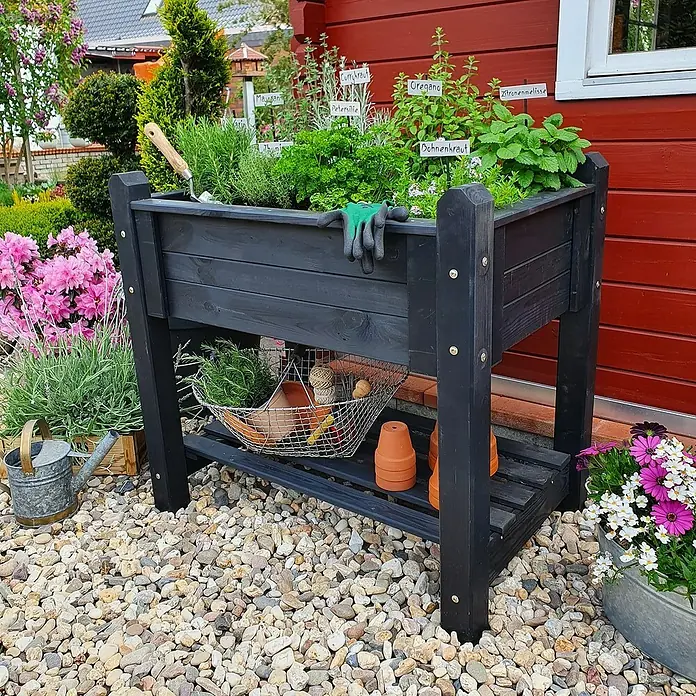 Promadino Kräuterhochbeet - L x B x H: 90 x 60 x 84,5 cm, Anthrazit - Nahaufnahme eines Hochbeets aus dunklem Holz mit Kräutern, Gartenwerkzeug und Dekorationselementen auf Kies.