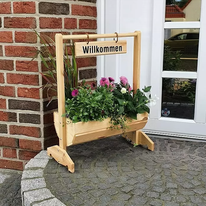 Promadino Pflanzkasten Willkommen - Außenmaß (L x B x H): 59 x 30 x 73 cm, Holz, Braun - Pflanzkasten aus Naturholz mit „Willkommen“-Schild, gefüllt mit bunten Blumen, neben einer Ziegelwand und weißen Eingangstür.
