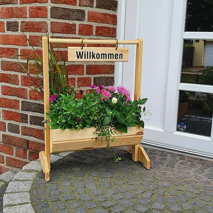 Promadino Pflanzkasten Willkommen - Außenmaß (L x B x H): 59 x 30 x 73 cm, Holz, Braun - Holzpflanzkasten mit der Aufschrift "Willkommen" und bunten Blumen, der vor einer Backsteinmauer und einer Tür steht.