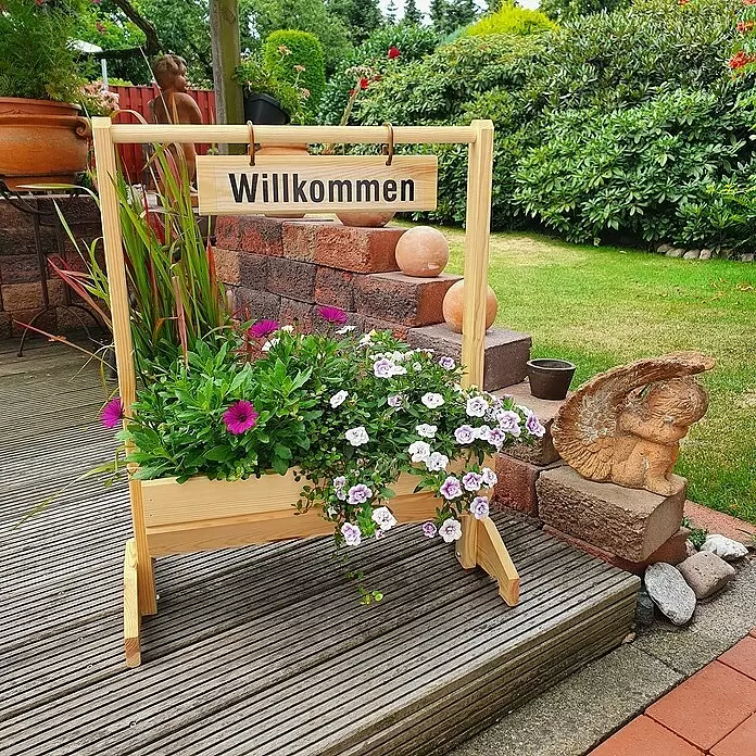 Promadino Pflanzkasten Willkommen - Außenmaß (L x B x H): 59 x 30 x 73 cm, Holz, Braun - Heller Holzpflanzkasten mit 'Willkommen'-Schild, gefüllt mit bunten lila, rosa und weißen Blumen, auf einer Holzterrasse im Garten.