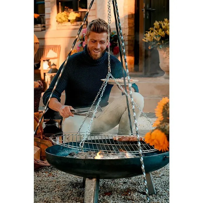 Buschbeck Schwenkgrill - Passend für: Buschbeck Feuerschale Ø 80 - 100 cm - Ein Mann grillt Fleisch auf einem Dreibein-Grill im Freien, während er eine Zange hält und lächelt.