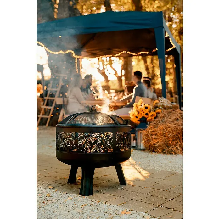 Buschbeck Feuerschale Leaves - Durchmesser: 63 cm, Stahl - Gemütliche Szene im Freien mit einem schwarzen Feuerkorb im Vordergrund, der Wärme und Atmosphäre für ein Abendessen im Freien bietet.