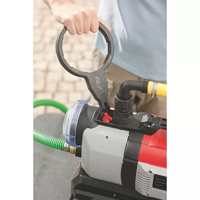 AL-KO Comfort Hauswasserautomat 4500 - 1.300 W, Max. Fördermenge: 4.500 l/h, Max. Druck: 5 bar - Eine Person verwendet einen schwarzen AL-KO Schraubenschlüssel, um das transparente Filtergehäuse einer rot-grauen Wasserpumpe zu bedienen.