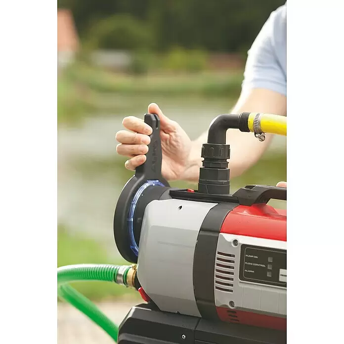 AL-KO Comfort Hauswasserautomat 4000 - 1.000 W, Max. Fördermenge: 4.000 l/h, Max. Druck: 4,5 bar - Person bedient eine rot, grau und schwarz lackierte Gartenpumpe mit grünen und gelben Schläuchen, im Freien an einem Gewässer.