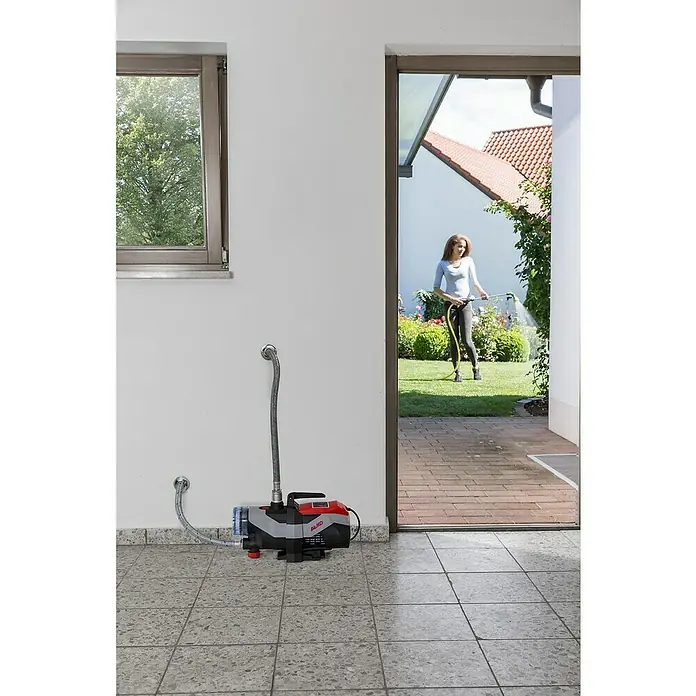 AL-KO Easy Gartenpumpe Jet 3600 Easy - 850 W, 3.600 l/h, 3,8 bar - AL-KO Gartenpumpe im Einsatz mit einer Frau, die im Garten mit einem Schlauch gießt, im Hintergrund ein Haus mit roten Ziegeln.