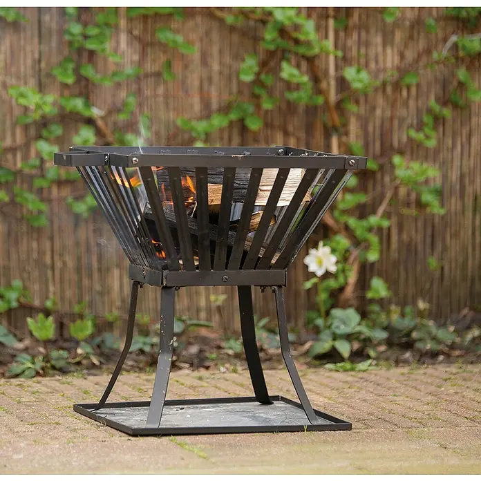 Feuerkorb Denver - L x B x H: 39 x 39 x 45 cm, Schwarz, Stahl - Der dunkle Feuerkorb ist mit Holz gefüllt, das bereits brennt. Er steht auf einem gepflasterten Untergrund mit einem Bambuszaun im Hintergrund.