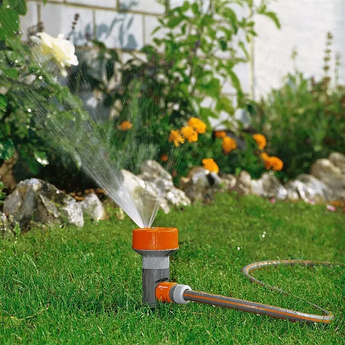 Gardena Classic Regner 4-Flächen-Regner TWIST - Max. Regnerfläche: 100 m², Anzahl Sprühformen: 4 Stk., Erdspieß - Ein Gardena Regner bewässert den Rasen, indem er Wasser versprüht. Das Gerät steht auf grünem Gras neben einem Gartenschlauch.