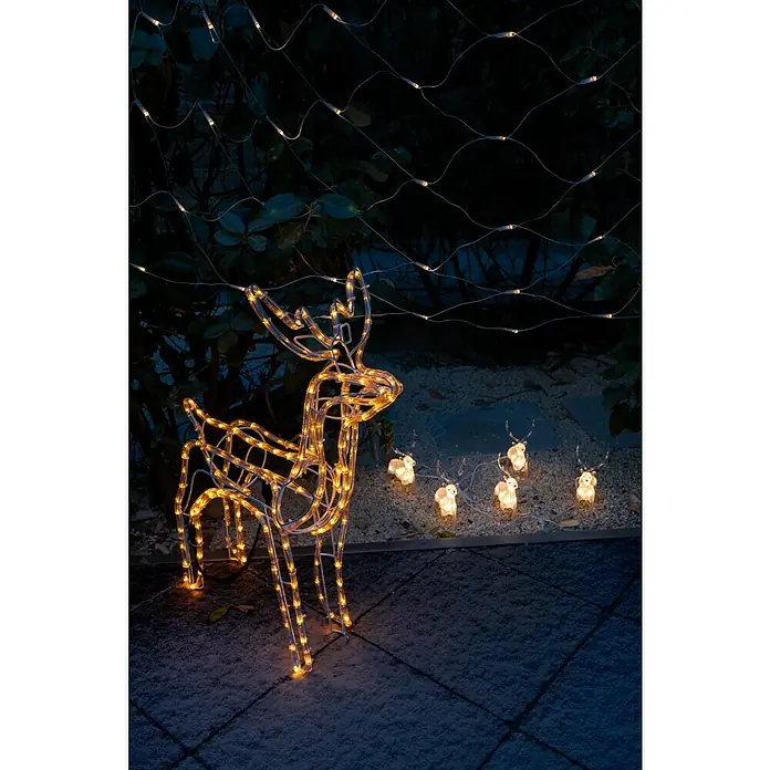 Konstsmide LED-Weihnachtsdekoleuchte LED Acryl - 4 m x 13 cm x 19 cm, Weiß, Netzbetrieben, LED - Beleuchtete Rentier-Dekoration mit kleineren Rentieren auf Pflastersteinen im Freien, umgeben von Pflanzen und Lichterketten.