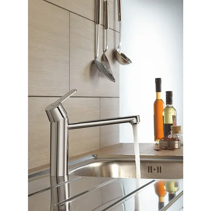 Grohe QuickFix Spültischarmatur Get - Chrom, Glänzend - Nahaufnahme eines glänzenden Wasserhahns mit fließendem Wasser in einer modernen Küche, Küchenutensilien und Flaschen im Hintergrund.