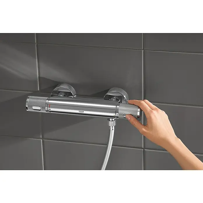 Grohe Precision Feel Thermostatarmatur - Breite: 31,3 cm, Chrom, Glänzend - Nahaufnahme einer Hand, die einen eleganten Grohe Thermostat mit Chromoberfläche vor grauen Fliesen bedient und die Temperatur einstellt.