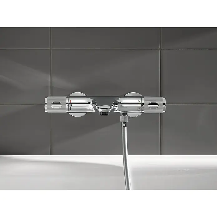 Grohe Precision Feel Wannenthermostat - Chrom, Glänzend - Nahaufnahme einer verchromten Duscharmatur mit Temperaturregler und Brauseschlauch vor grauen Fliesen.