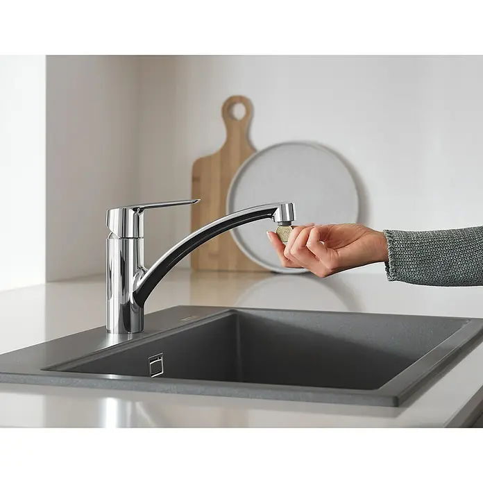 Grohe QuickFix Spültischarmatur Start - Ausladung Armatur: 227 mm, Chrom, Schwenkbereich: 140 ° - Glänzende Küchenarmatur mit einer Hand, die eine Münze hält, vor einem grauen Spülbecken und neutralem Hintergrund.