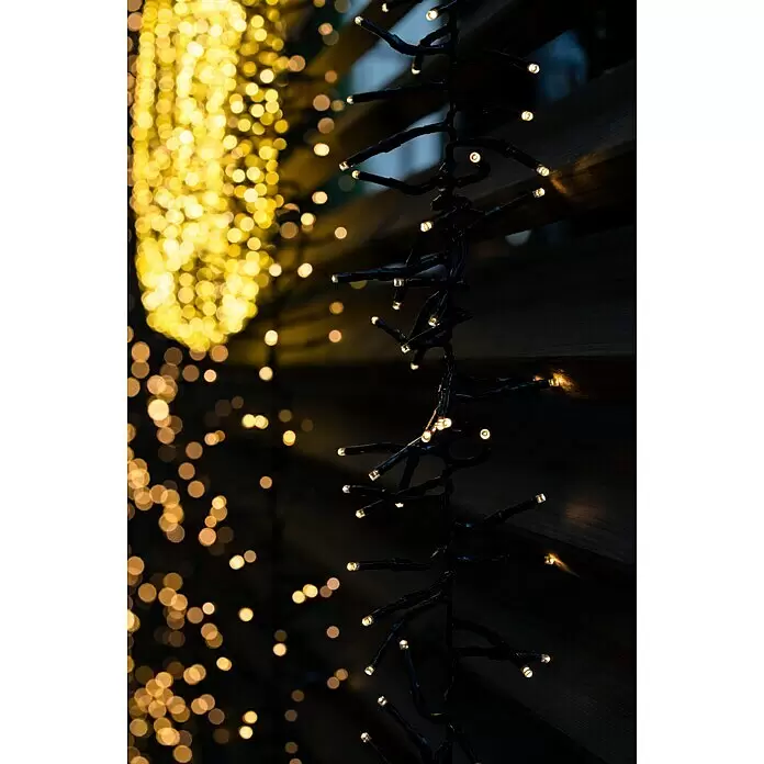 Tween Light LED-Clusterlichterkette - Außen, Länge: 8,15 m, 768 Stk., Netzbetrieben, Lichtfarbe: Warmweiß - Dekorative Lichterkette mit warmweißen LEDs an schwarzem Kabel, unscharfe Lichter im Hintergrund.