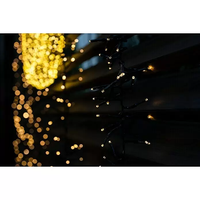 Tween Light LED-Clusterlichterkette - Außen, Länge: 8,15 m, 768 Stk., Netzbetrieben, Lichtfarbe: Warmweiß - Warmweiße LED-Lichterkette beleuchtet eine dunkle Struktur mit goldenen Bokeh-Lichtern im unscharfen Hintergrund.