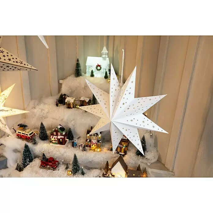 Markslöjd LED-Stern Saturnus - 45 cm, Weiß, IP20, Netzbetrieben - Weiße, sternförmige Papierlaternen mit ausgestanzten Sternen und Punkten, die über einem festlichen Miniatur-Weihnachtsdorf hängen.