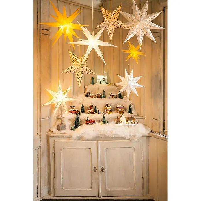 Markslöjd LED-Stern Gisel - 45 cm, Silber, Weiß, IP20, Netzbetrieben - Festliche Weihnachtsszene mit leuchtenden Sternen und Miniaturdorf auf einer Kommode.