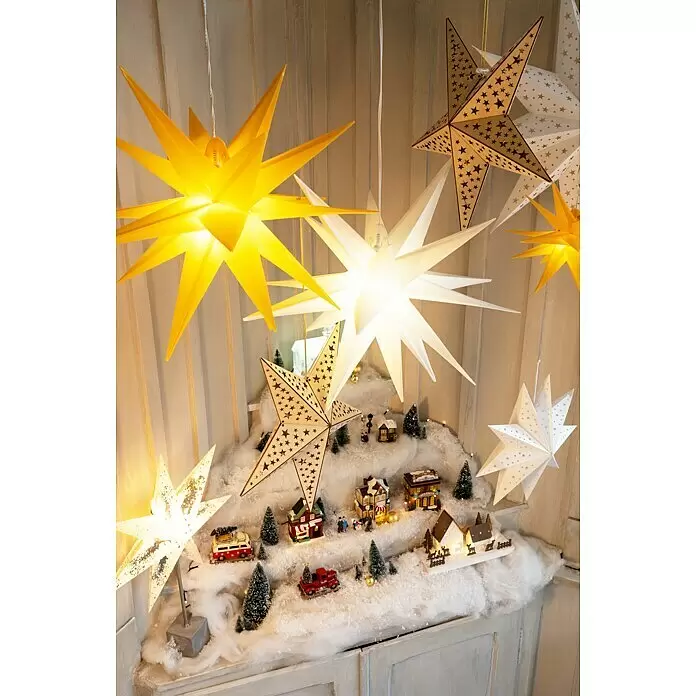 Markslöjd LED-Stern Gisel - 45 cm, Silber, Weiß, IP20, Netzbetrieben - Verschiedene beleuchtete Sternlaternen, darunter gelbe, weiße und hölzerne, über einer Weihnachtsdorf-Szene hängend.