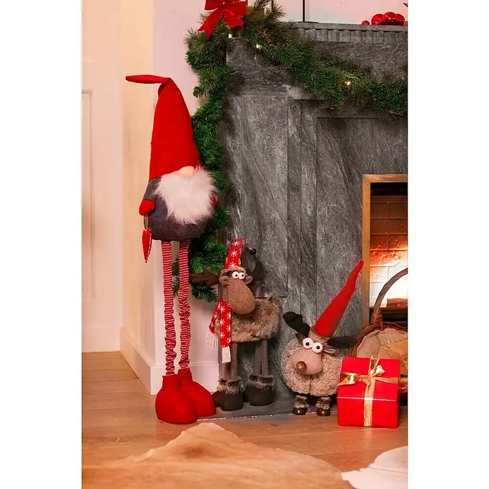 Dekofigur Elch mit Mütze - L x B x H: 35 x 15 x 53 cm, Kunststoff, Rentier - Weihnachtsdekoration mit einem großen roten Wichtel, zwei Elchen und einem roten Geschenk vor einem Kamin.