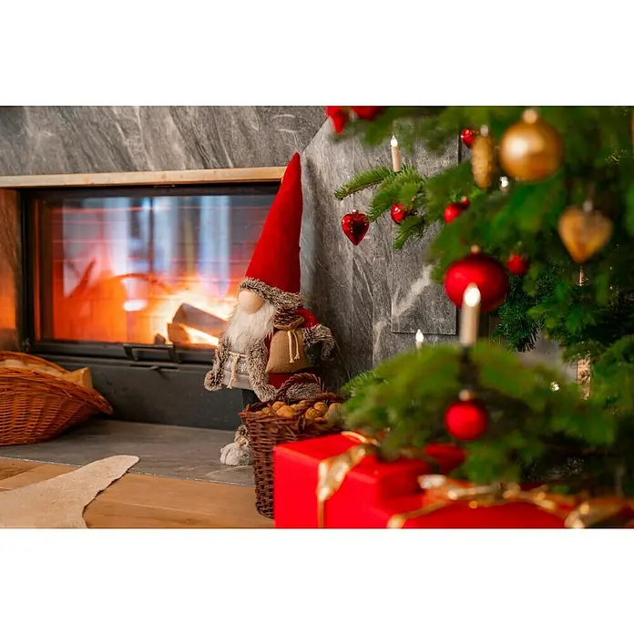 Dekofigur wackelnder Wichtel mit Jutebeutel - L x B x H: 24 x 19 x 78 cm, Textil - Gemütliche Weihnachtsszene mit einem festlichen Wichtel neben einem Kamin und einem geschmückten Weihnachtsbaum mit roten und goldenen Kugeln.
