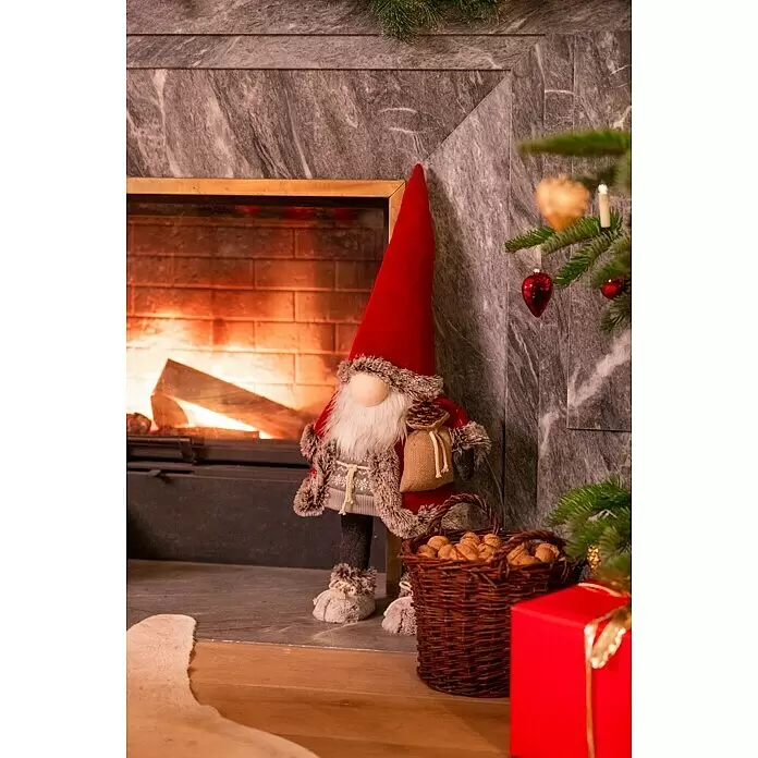 Dekofigur wackelnder Wichtel mit Jutebeutel - L x B x H: 24 x 19 x 78 cm, Textil - Ein festlicher Weihnachtswichtel mit roter Mütze, weißem Bart und Pelzbesatz steht an einem Kamin und hält einen Sack und einen Tannenzapfen.
