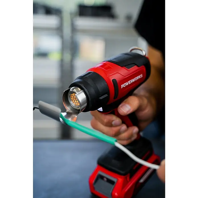Powerworks Akku-Heißluftgebläse P24HG - 24 V, Ohne Akku, Temperaturbereich: 550 °C - Ein rotes Powerworks-Heißluftgebläse erhitzt einen Schrumpfschlauch an einem Kabel. Das Werkzeug liegt gut in der Hand.