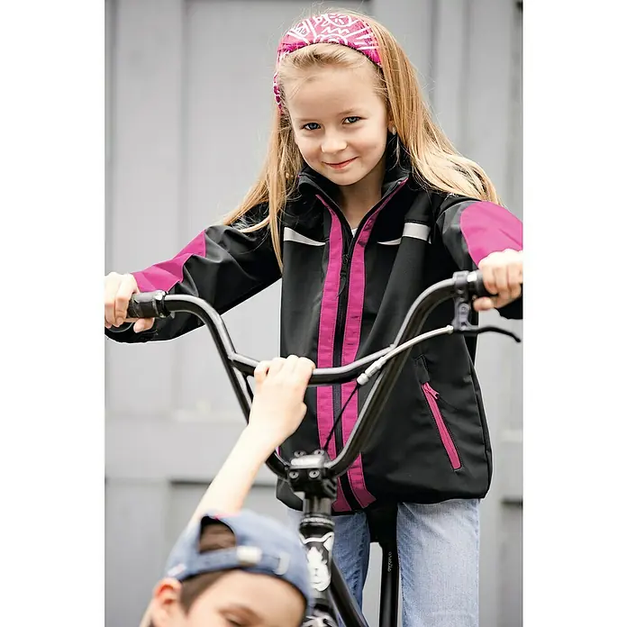 Kübler Softshell-Jacke Kidz110, Schwarz/Pink - Ein Mädchen mit einem pinkfarbenen Stirnband und einer schwarz-pinken Jacke auf einem Fahrrad.