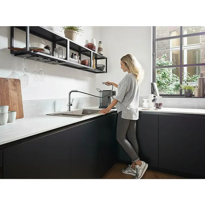 Hansgrohe Spültischarmatur Talis M54 270Chrom, Glänzend - Eine Frau befüllt mit einem ausziehbaren Küchenarmatur einen Behälter in einer modernen Küche mit dunklen Unterschränken und Regalen.