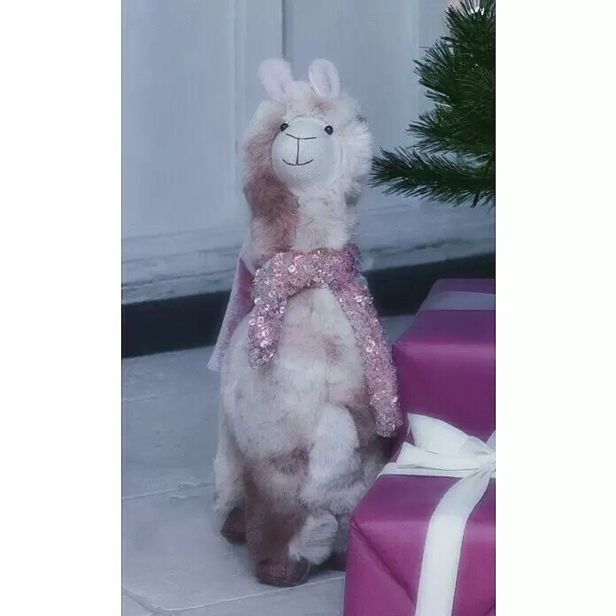 Dekofigur Weihnachtslama - L x B x H: 23 x 13 x 42 cm, Textil, Weihnachtswichtel, Schneemann - Ein flauschiges rosa-braunes Lama-Plüschtier mit Paillettenschal sitzt neben lila Geschenken und einem Weihnachtsbaum.