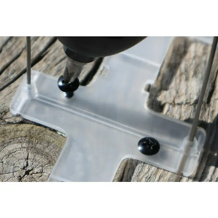 Altadex Sistema antipalomas - Largo: 200 cm, Acero - Primer plano de un destornillador atornillando un tornillo negro en una placa de plástico transparente en forma de cruz, colocada sobre una superficie de madera.
