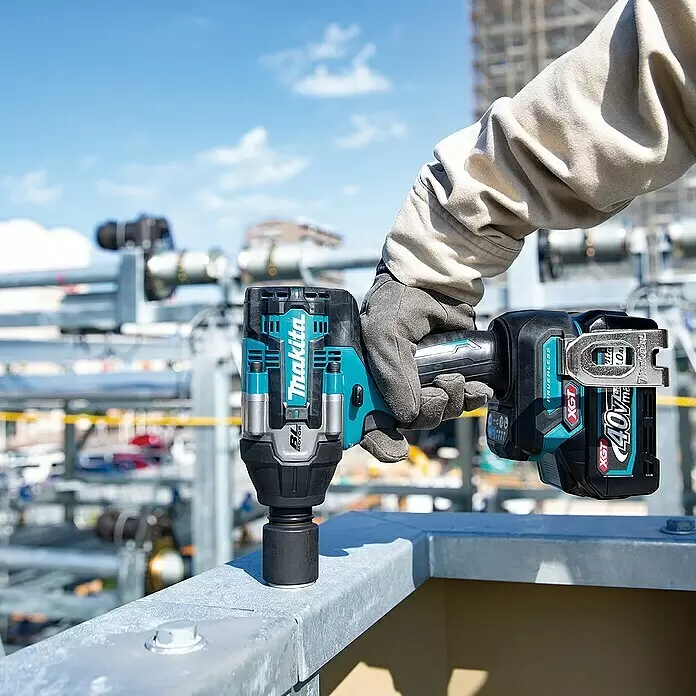 Makita XGT 40V max. Akku-Schlagschrauber TW008GZ - 40 V, Ohne Akku, 760 Nm - Eine behandschuhte Hand hält einen blau-schwarzen Makita XGT 40V Schlagschrauber mit Stecknuss auf einem Metallgeländer einer Baustelle.