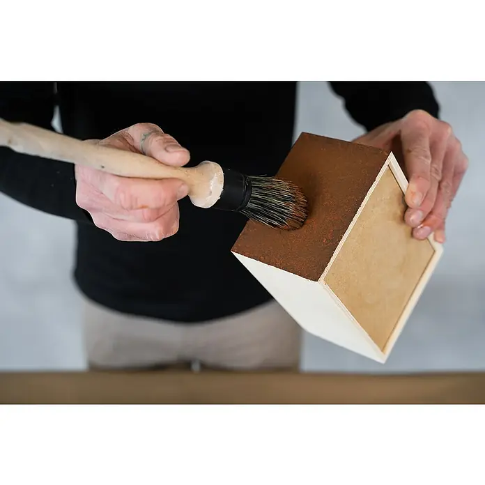 Montó Bricolovers Yeso con efecto de color - Óxido, 2,5 l, Mate - Primer plano de una persona pintando una caja de madera con un pincel y pintura marrón. La caja está casi terminada de pintar.