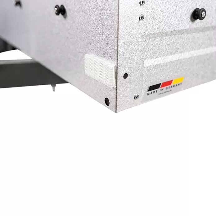 Stema Anhänger Mini 350 mit Bordwandaufsatz u. Flachplane - Nutzlast: 245 kg, Einachser, Kasteninnenmaß: 134 x 108 x 26,5 cm (Höhe inkl. Bordwandaufsatz 53 cm), Ungebremst - Ecke eines silber-grauen, texturierten Metallkastens mit weißem Reflektor und "Made in Germany" Etikett.