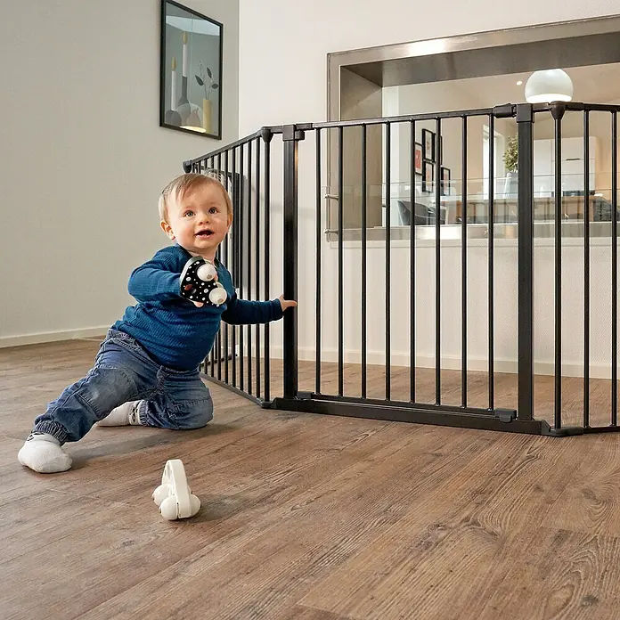 Star Stairs Sicherheitsgitter Ben - L x H: 278 x 71 cm, Geeignet für: Kinder bis 24 Monate - Ein niedliches Kind spielt vor einem schwarzen Treppenschutzgitter im Haus, das Sicherheit bietet und den Zugang zu Treppen oder anderen Bereichen einschränkt.