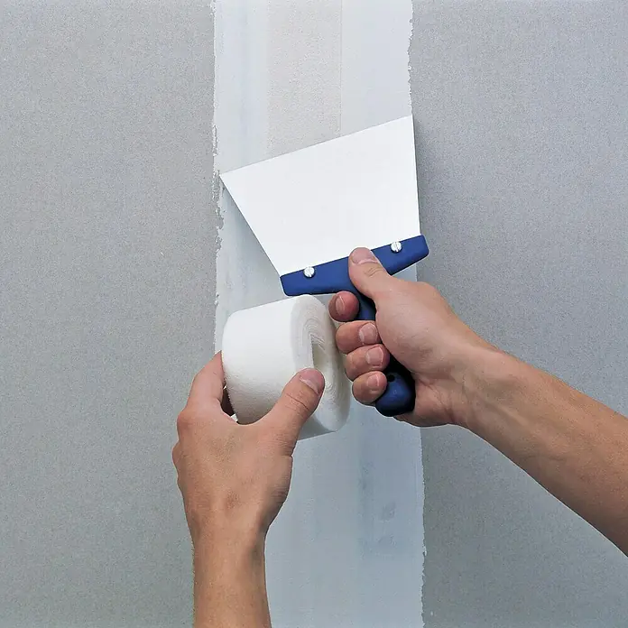 Rigips Gitterbewehrungsstreifen - 90 m x 48 mm - Nahaufnahme einer Person, die mit einem Spachtel und Klebeband eine Wand verputzt, um eine glatte Oberfläche zu schaffen.