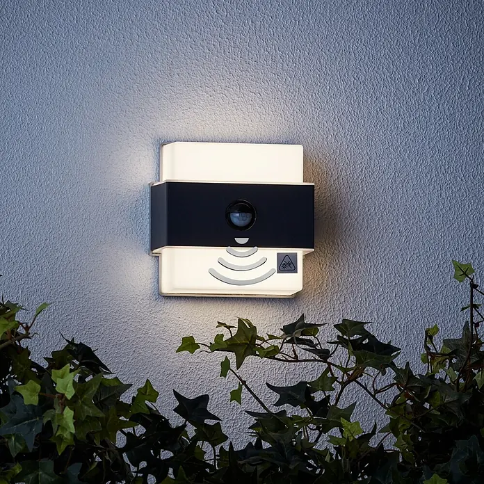 Brilliant LED-Sensor-Außenwandleuchte Archie - 7,5 W, L x B x H: 7 x 14,5 x 15 cm, Anthrazit, Neutralweiß - Moderne Außenleuchte mit Bewegungsmelder und integrierter Beleuchtung an einer strukturierten Wand, umgeben von Grün.