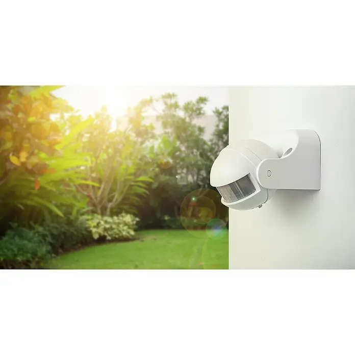 Garza Sensor de movimiento de paredes - Plástico, Blanco - Primer plano de un sensor de movimiento blanco montado en una pared blanca, con vistas a un jardín al fondo.