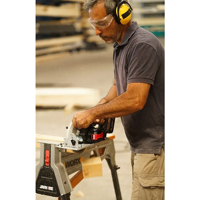 Worx PowerShare 20V Akku-Handkreissäge WX520.9 - 20 V, Ohne Akku, Durchmesser Scheibe: 185 cm - Ein Mann sägt ein Holzbrett mit einer WORX Kreissäge, während er Schutzbrille und Ohrenschützer trägt.