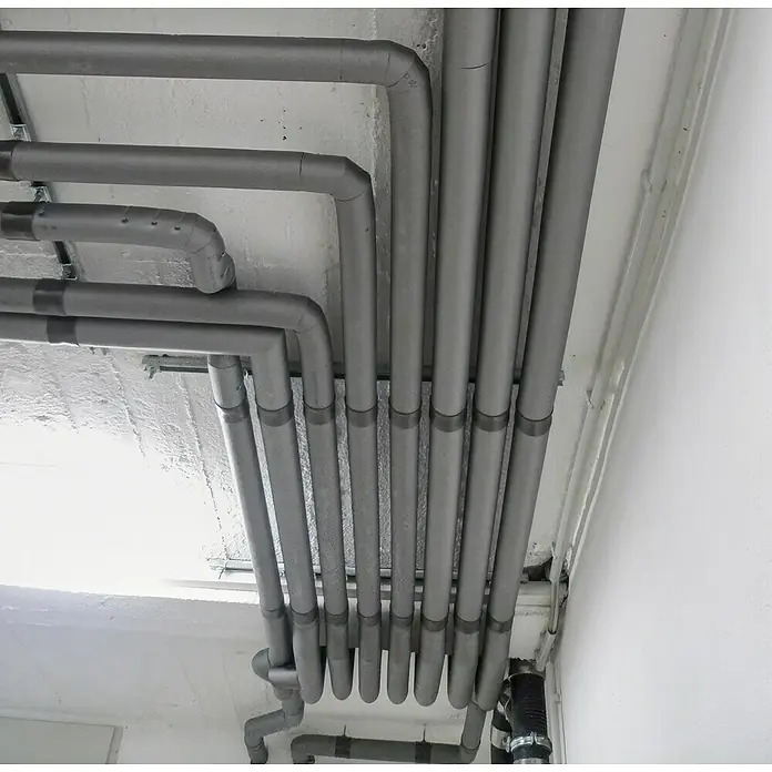 Thermaflex ThermaGo PE-Rohrisolierung 35/20 - Durchmesser: 35 mm, Dämmstärke: 20 mm, Selbstklebend, Länge: 1 m - Graue Rohre verlaufen unter einer Decke und entlang einer Wand. Isolierte Rohre sind mit dunklen Manschetten versehen.