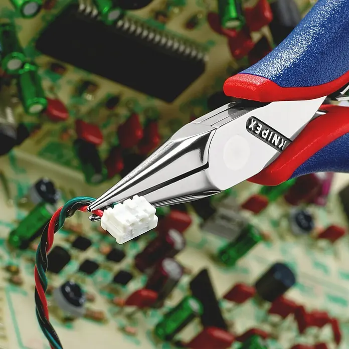 Knipex Flachrundzange - Länge: 11,5 cm, Backenform: Flachrund - Knipex Präzisions-Elektronikzange hält einen weißen Stecker mit Kabeln vor einer Platine. Ideal für filigrane Arbeiten in der Elektronik.