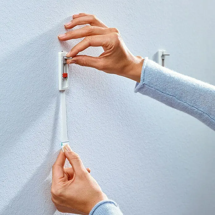 Tesa Tornillo adhesivo ajustable - Rectangular, Carga soportada: 1 kg, 2 ud. - Una persona fija una tira adhesiva blanca con ganchos en una pared blanca para colgar algo.