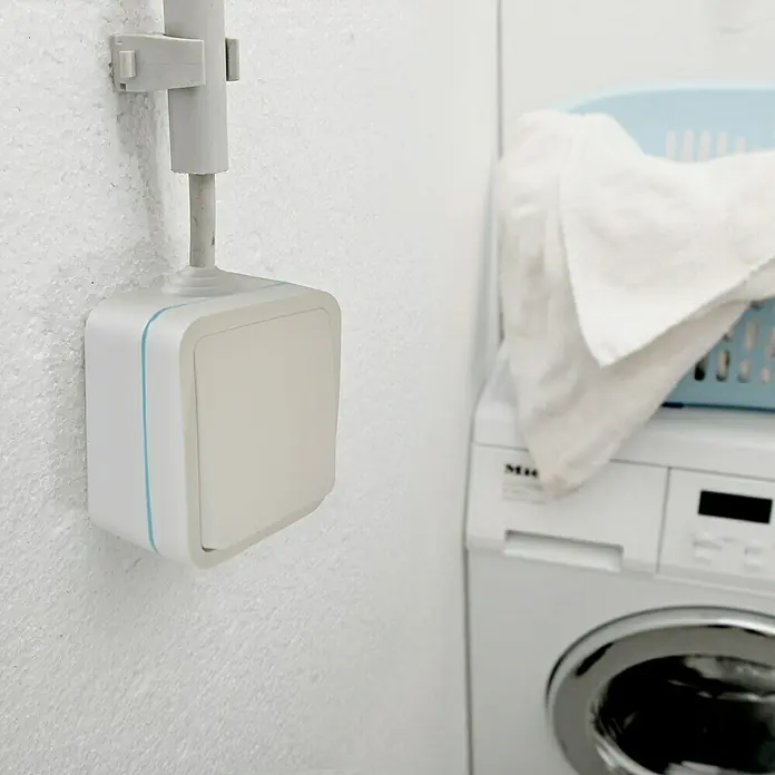 Voltomat Blueline Feuchtraum-Wechselschalter - Weiß, Aufputzmontage, IP54 - Nahaufnahme eines Smart Home Schalters mit blauen Akzenten, der an einer weißen Wand montiert ist. Waschmaschine im Hintergrund.