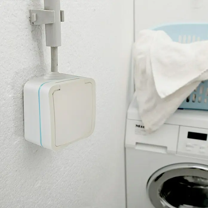 Voltomat Blueline Feuchtraum-Steckdose - 1-fach, Horizontal, IP54, Weiß - Eine Aufputzsteckdose mit blauem Akzent befindet sich an einer weißen Wand, neben einer Waschmaschine und einem Wäschekorb.