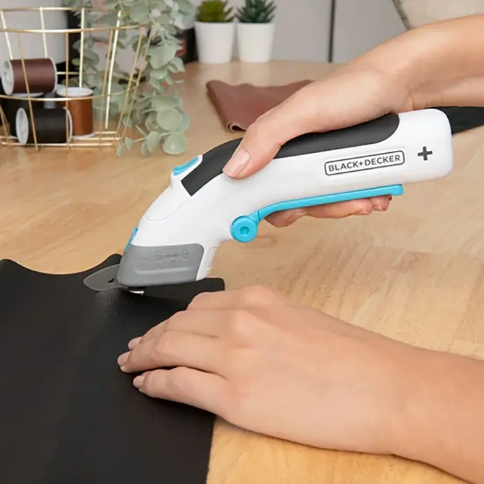 Black+Decker Tijera de batería BCSC115 3,6V-Li - 3,6 V, 1 batería, 1,5 Ah - Una persona está cortando un trozo de cuero negro sobre una mesa de madera con un cúter rotatorio Black+Decker.
