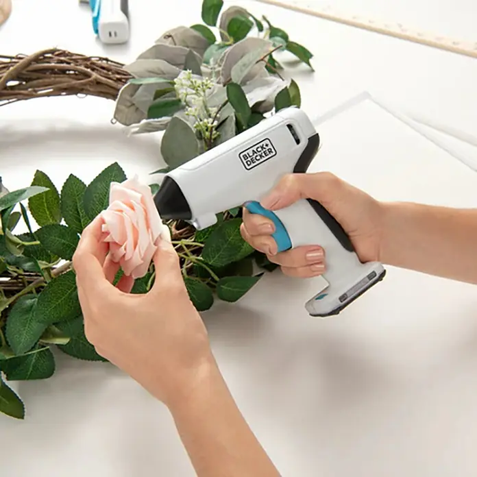 Black+Decker Pistola termoencoladora con batería BCGL115 3,6V-Li - 3,6 V - Primer plano de una persona que usa una pistola de pegamento caliente Black+Decker para pegar una rosa rosa a una corona.
