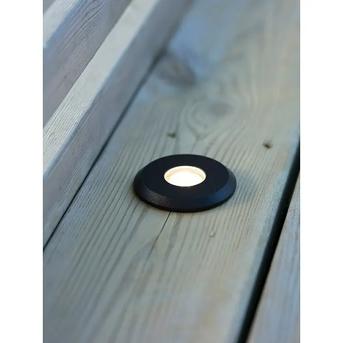 Garden 24 LED-Einbauspot - Warmweiß, Schwarz, 6 x 6 x 9,7 cm - Bodeneinbauleuchte auf einer Holzterrasse. Die Leuchte ist rund, schwarz und strahlt ein warmes, helles Licht aus.
