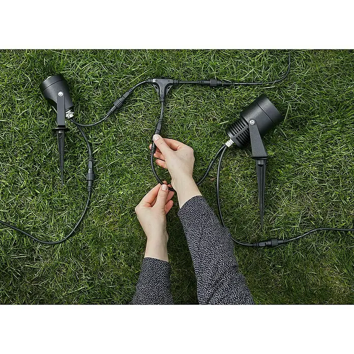 Garden 24 Verteiler Kabel - 26 x 26 cm - Gartenstrahler auf Rasen mit Händen, die die Kabel verbinden. Die Strahler sind schwarz und haben Erdspieße.