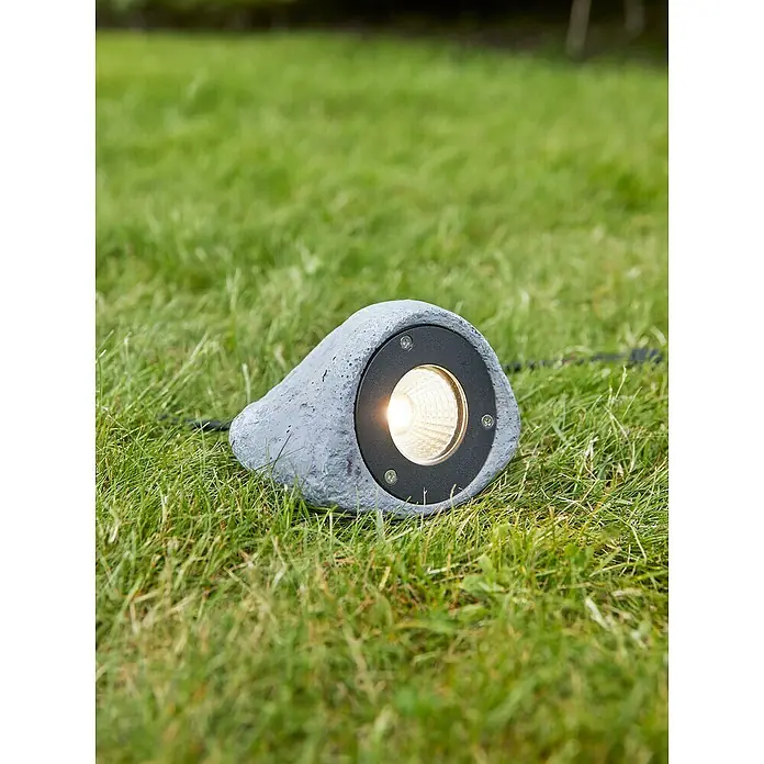 Garden 24 LED-Gartenspot - 3 W, 11 cm - Eine graue Gartenleuchte in Steinoptik sitzt im grünen Gras und strahlt helles Licht aus. Perfekt für stimmungsvolle Beleuchtung im Außenbereich.